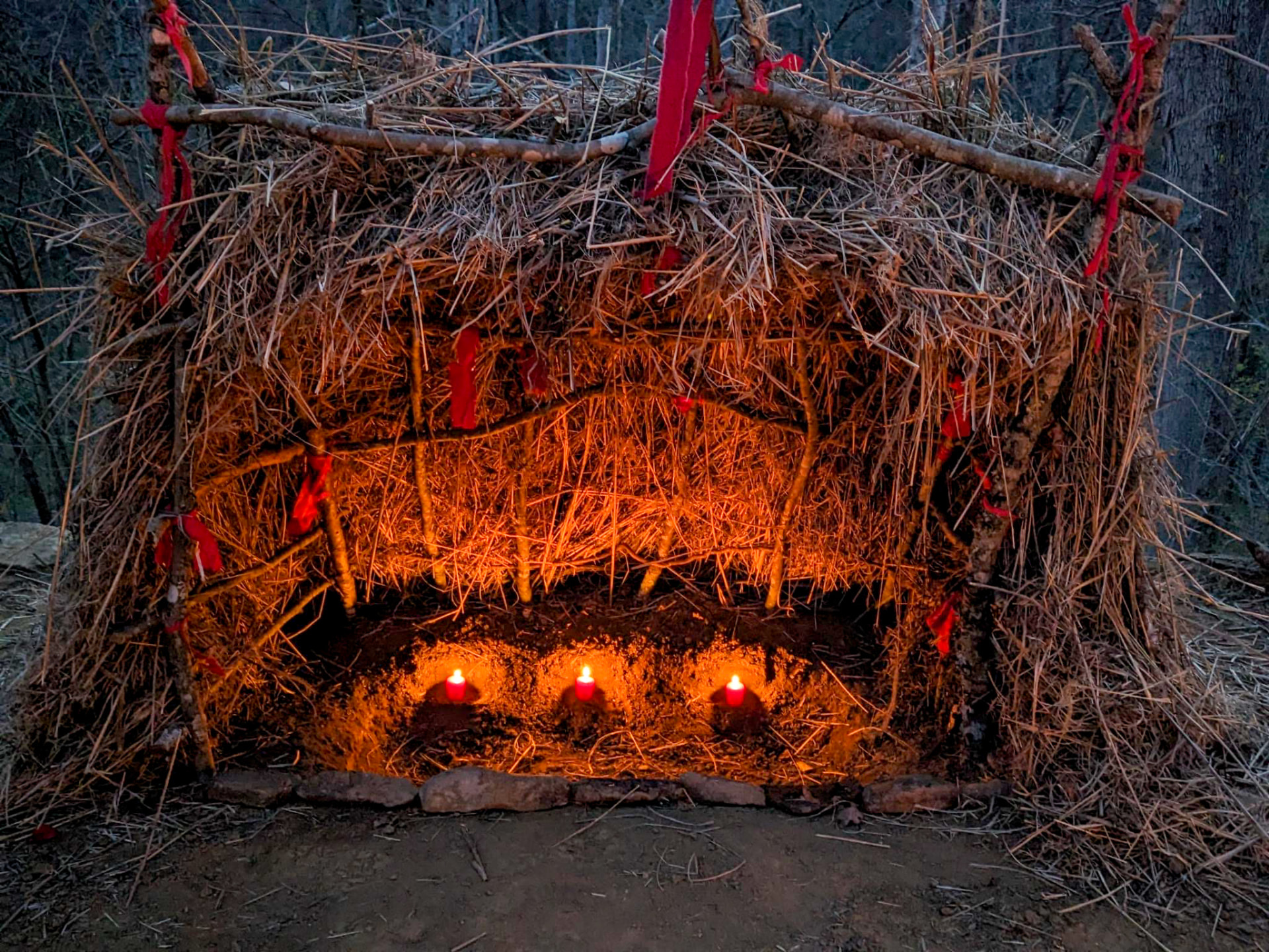 A straw threshold shelter lit from within by three red candles at dusk
